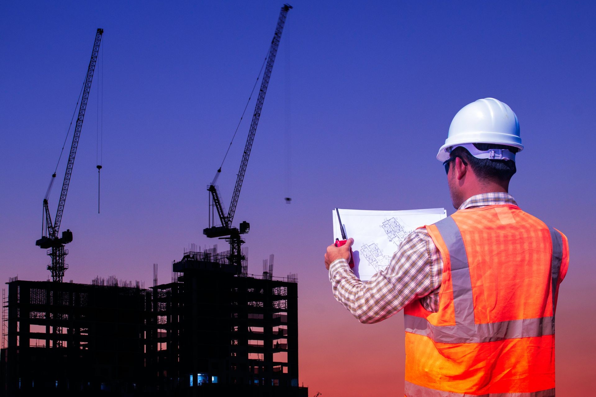 Engineer with blue print for control working at large building construction site in sunset